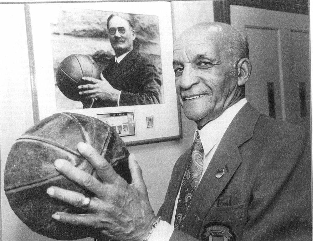 A smiling man holds up a basketball, standing in front of a framed photo of James Naismith in the same pose.