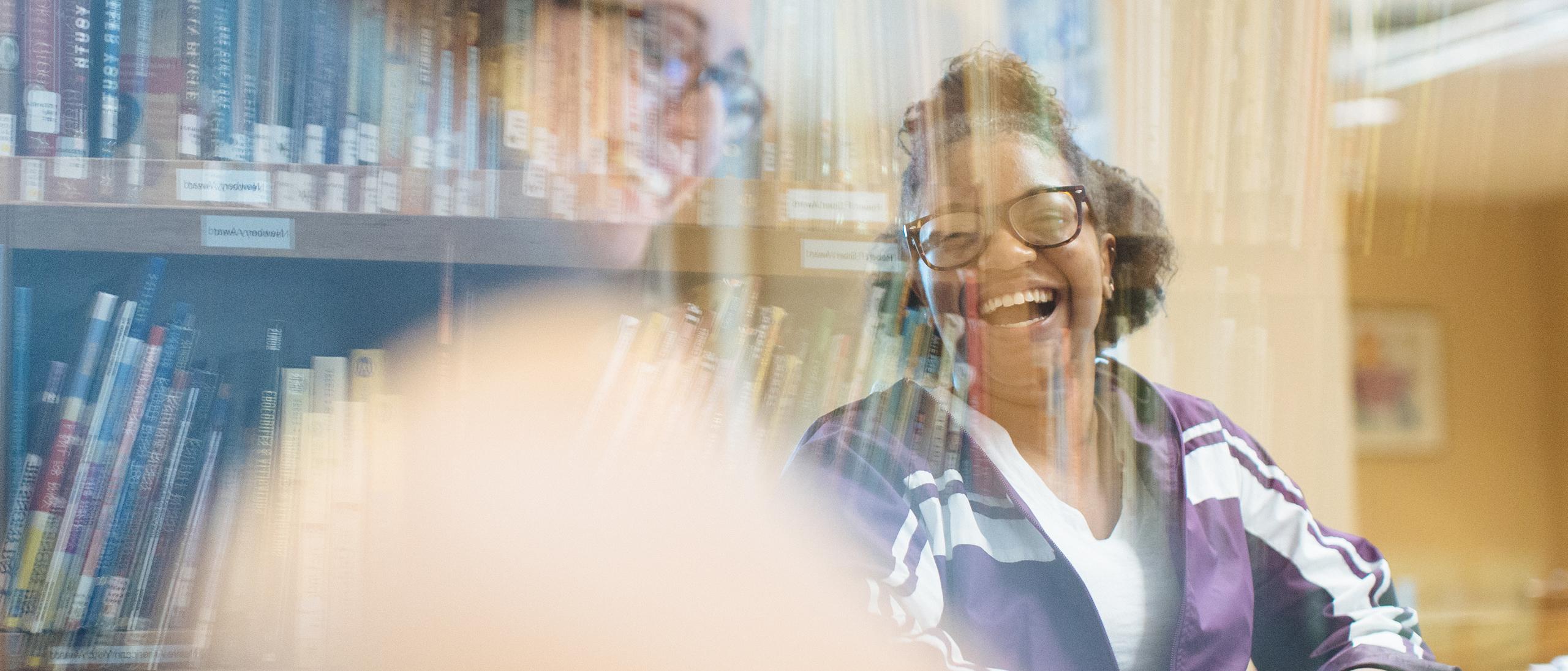 Two smiling students read and review children's literature with book shelves in the background