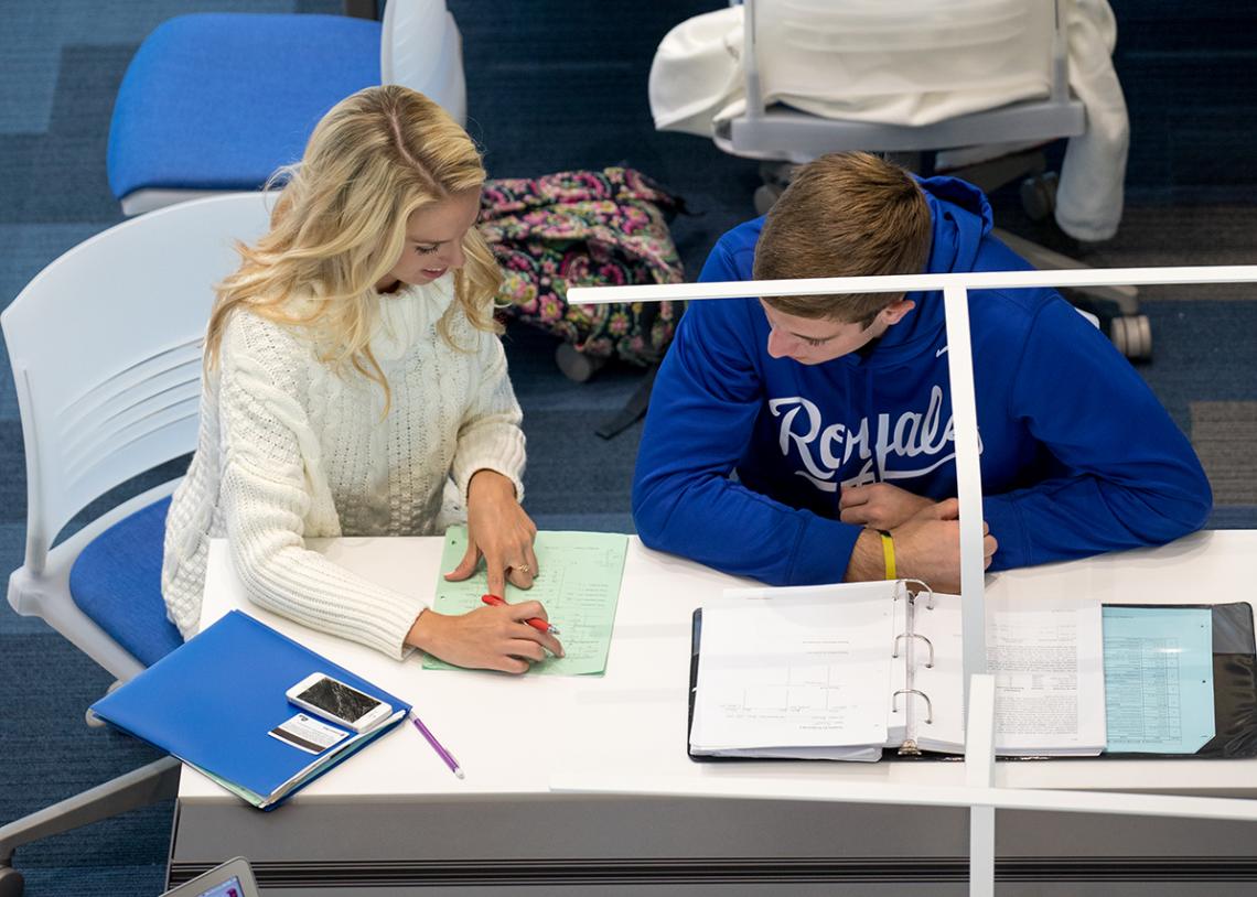 Two students are working together at a table with open notebooks